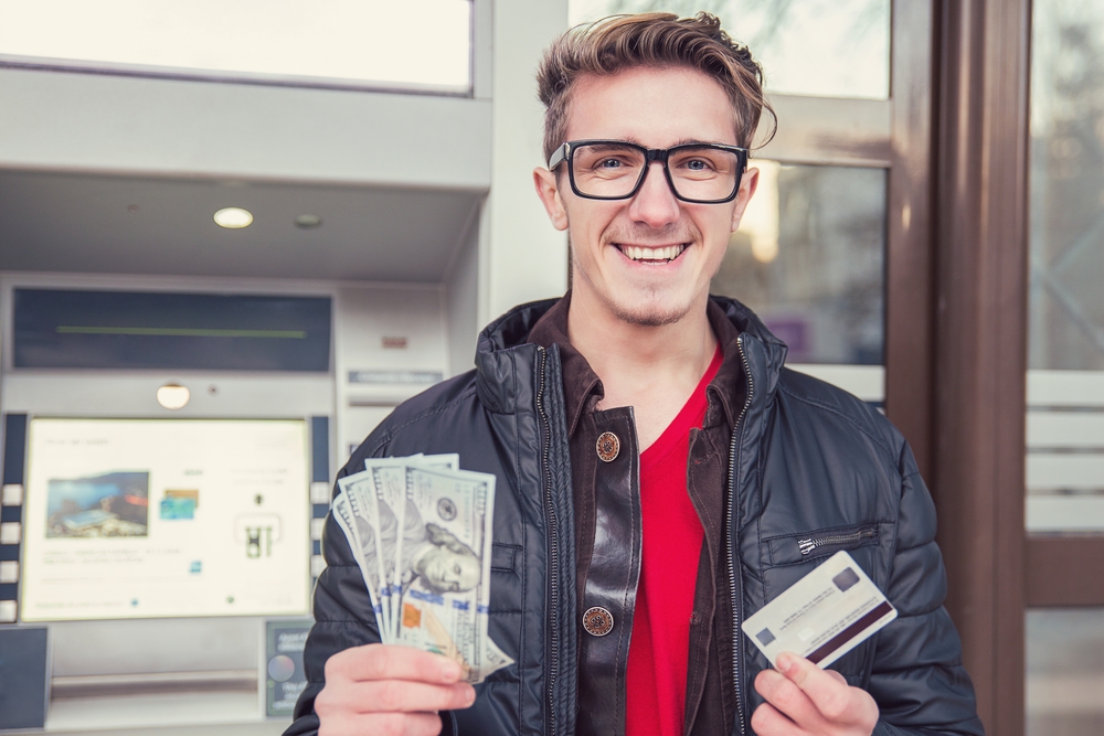 Choosing the Right Credit Card: Happy young man showing credit card and cash while standing near an ATM machine 