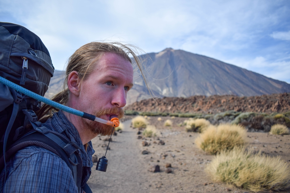 What Not to Do When Hiking: Pretty young traveler man hiking drinking water on mountain with Teide volcano on the background. Enjoying hot weather and sun in Spain. Staying hydrated on sport hike. Hydration bladder. 