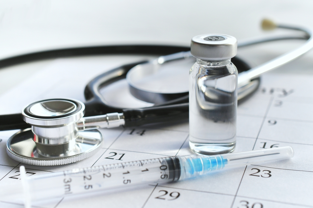 The Worst Places to Visit Without Travel Vaccinations: A medical syringe needle with a plain unmarked vial of medicine laying on a calendar page with a stethoscope. 