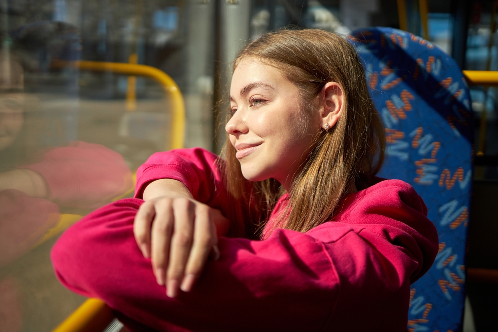 A Visitor's Guide to Getting Around Lisbon’s Trams: Young girl in pink hoodie sitting in public transport, modern tram, bus, looking out the window and smiling. 