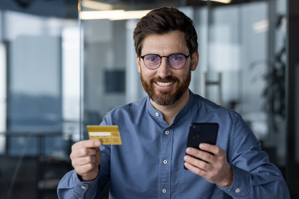 Managing Cash or Cards Money Abroad: Close-up portrait of a young man in glasses and shirt sitting in a modern office at the workplace, holding credit card and mobile phone, smiling and looking at camera. 