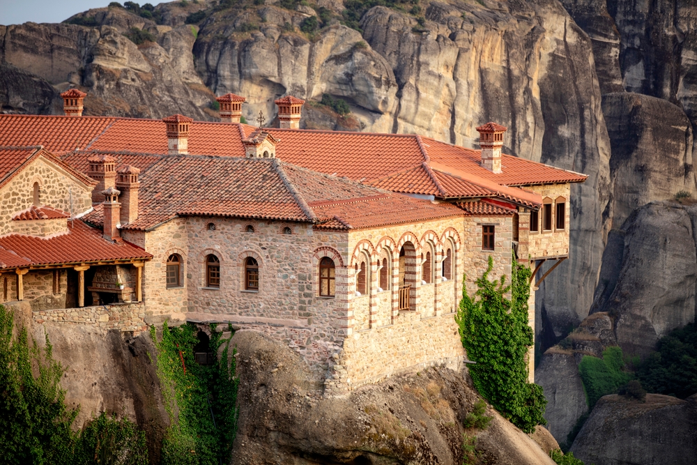 Sustainable Travel in Greece: Aerial view of Meteora Monastery complex on rocks, illuminated by rising sun against mountains in background. 