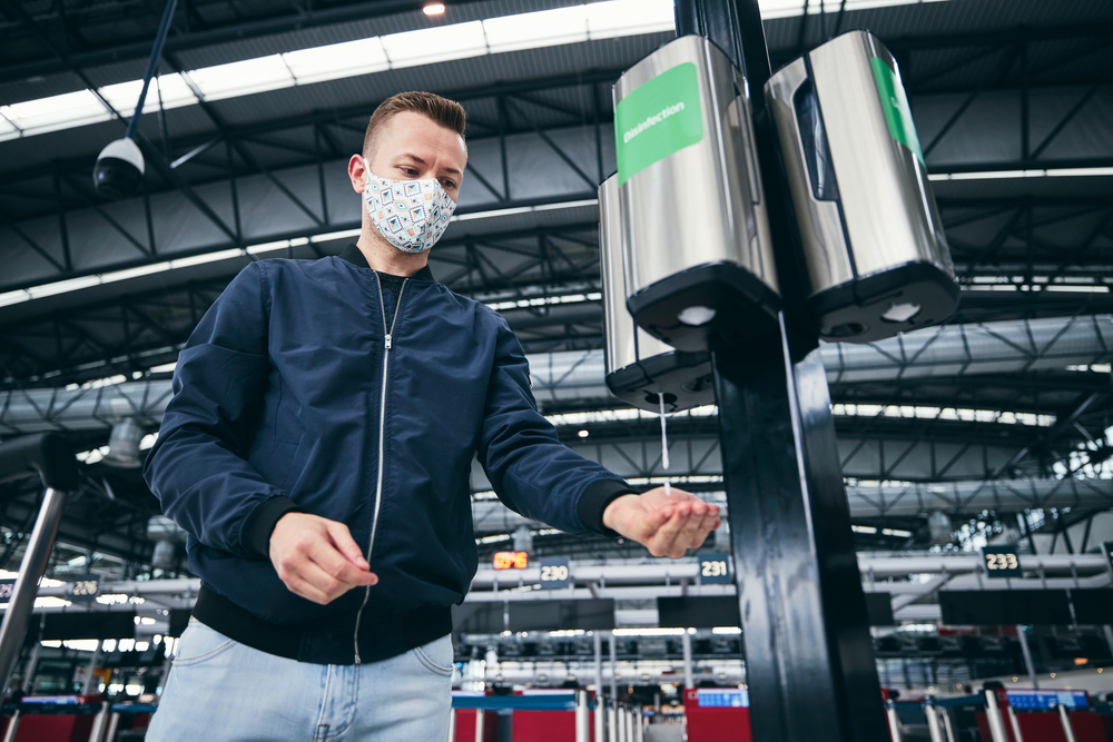 Breezing through Security and Layovers: Man wearing face mask and using hand sanitizer at airport. Themes traveling during pandemic, hygiene and personal protection. 