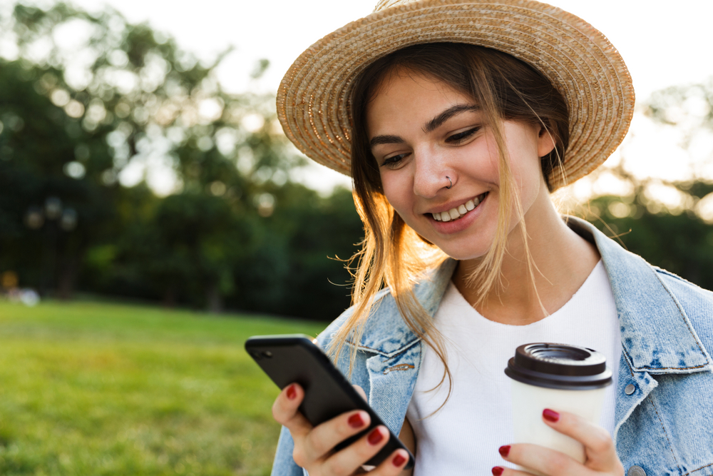 How to Stay Connected Abroad Without Breaking the Bank: Lovely young girl in summer hat sitting outdoors, using mobile phone, holding cup of coffee 