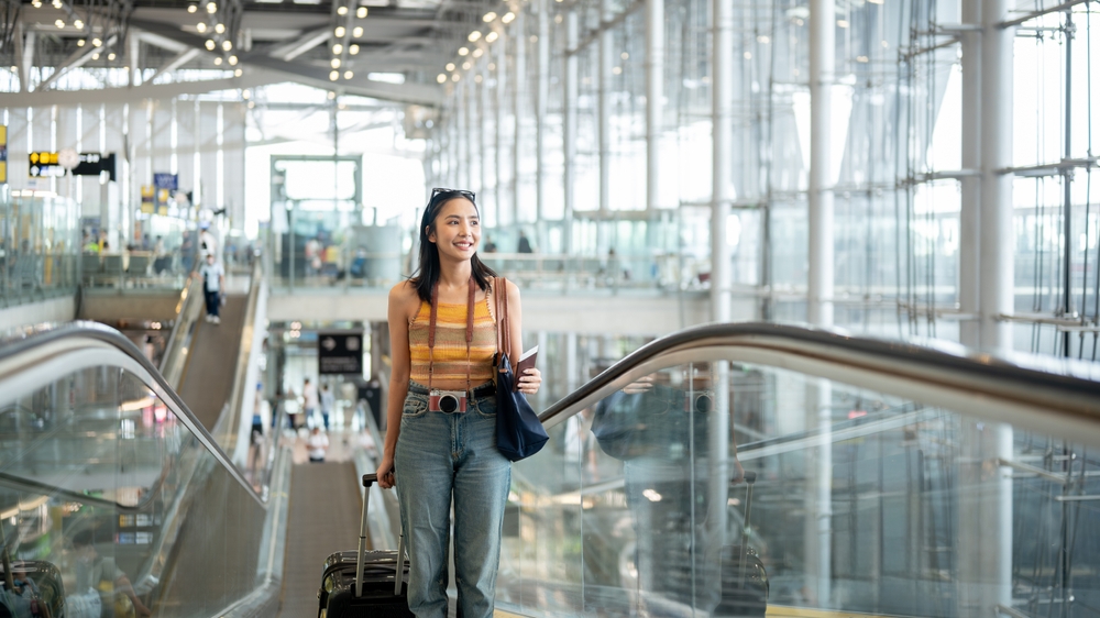 Tips for Finding Cheap Flights: A Step-by-Step Guide: A gorgeous young Asian female tourist passenger is on an airport escalator, carrying her luggage, going to the airport check-in counter, traveling by plane. people and transportation concepts 