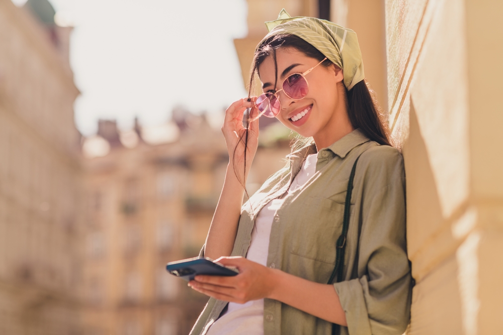 How to Choose the Best Accommodation for Your Travel Style: Portrait of attractive cheerful lady toothy smile hand touch sunglass hold telephone walk city center outside