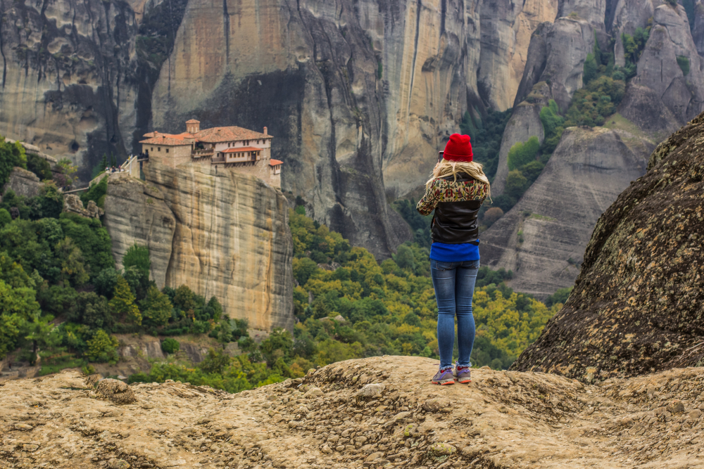 The Worst Times to Visit Meteora: Travel girl stay back to camera in autumn season clothes and bright red hat and taking mobile photo by the beautiful Greek European highland landscape with orthodox religion monastery on mountain top 