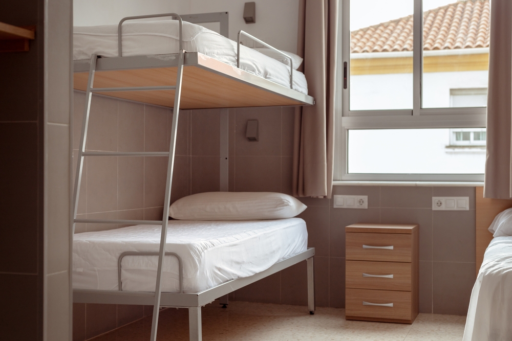 Most of Shared Hostel Accommodations: A clean and minimalistic hostel room with a bunk bed, white bedding, and a small wooden bedside table, ideal for budget travelers seeking affordable accommodation. 